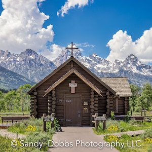 May include: A wooden log cabin church with a cross on the roof, set against a backdrop of snow-capped mountains and a blue sky with white clouds. The church has a wooden door and windows, and a sign that reads "Church of the  Transfiguration".