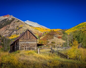 May include: A rustic wooden cabin sits in a valley surrounded by colorful autumn foliage and snow-capped mountains. The cabin has a wooden fence and a blue sky above.