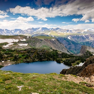 May include: A scenic view of a mountain lake surrounded by green hills and snow-capped peaks. The sky is blue with white clouds.