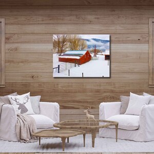 May include: A red barn with a white roof sits on a snowy field with trees in the background. The barn is surrounded by a fence and there is a small wooden building in the distance.