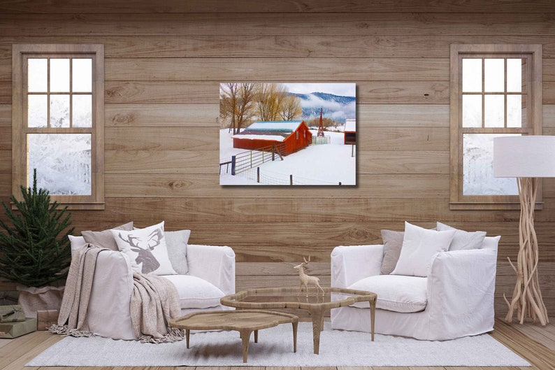 May include: A red barn with a white roof sits on a snowy field with trees in the background. The barn is surrounded by a fence and there is a small wooden building in the distance.