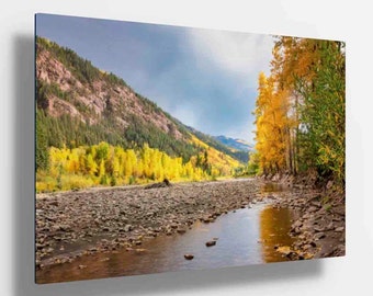 Op de afbeelding: Een landschapsfoto met een kronkelende rivier die door een vallei stroomt met een bergketen op de achtergrond. De bomen langs de rivier zijn in levendige herfstkleuren, met tinten geel, oranje en rood. De lucht is bewolkt met een vleugje blauw.