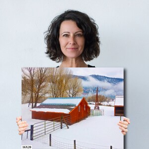 May include: A red barn with a white roof and a snow-covered field in front of it. The barn is surrounded by a wooden fence and there are trees in the background. The text "16X20" is printed in the bottom left corner of the image.