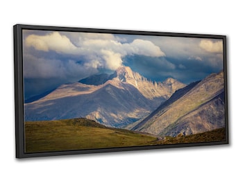 May include: A framed print of a mountain landscape. The image depicts a winding road through a valley, with mountains in shades of brown, green, and grey. The sky is filled with dark clouds and patches of sunlight, creating a dramatic effect.