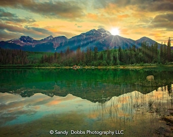 Puede incluir: Una fotografía de paisaje que representa una cordillera reflejada en un lago tranquilo al atardecer. El cielo es una mezcla vibrante de tonos naranja, rosa y morado, con el sol brillando a través de las nubes. Las montañas son de un azul profundo y el lago es de un verde profundo.