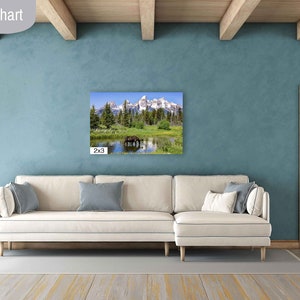 Op de afbeelding: Een witte bank met blauwe kussens in een woonkamer met een groot canvasprint van een berglandschap erboven. De canvasprint is in de linkerbenedenhoek met "2x3" gelabeld.