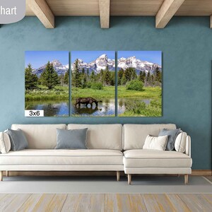 Op de afbeelding: Een driedelig canvasprint met een schilderachtig landschap met bergen, bomen en een meer. Een bruine eland drinkt uit het meer in het middelste paneel. De print is tentoongesteld op een witte bank in een woonkamersetting. De tekst "3x6" is zichtbaar in de linkerbenedenhoek van de afbeelding.