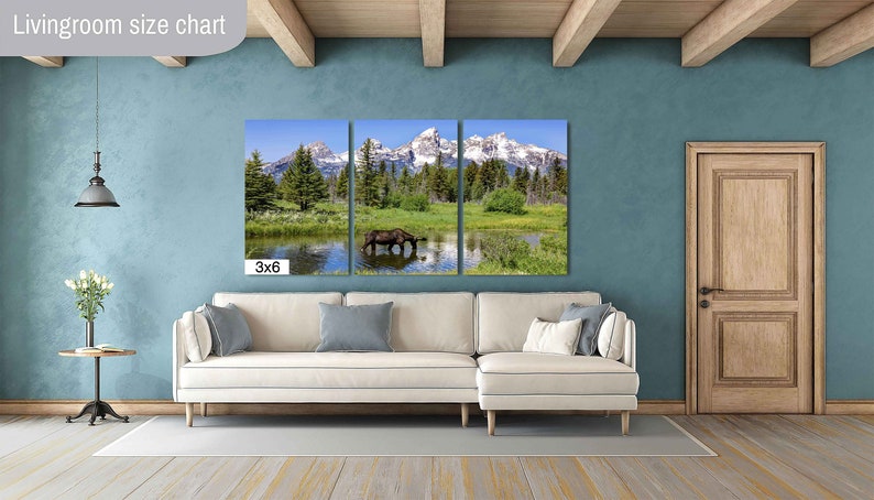 Op de afbeelding: Een driedelig canvasprint met een schilderachtig landschap met bergen, bomen en een meer. Een bruine eland drinkt uit het meer in het middelste paneel. De print is tentoongesteld op een witte bank in een woonkamersetting. De tekst "3x6" is zichtbaar in de linkerbenedenhoek van de afbeelding.