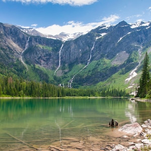 May include: A scenic landscape featuring a clear, turquoise lake reflecting the surrounding mountains and sky. Tall waterfalls cascade down the rocky cliffs, and evergreen trees line the shores. The image captures a serene natural environment.