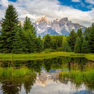 May include: A scenic view of a mountain range reflected in a still pond. The mountains are snow-capped and the pond is surrounded by lush green trees and grass.