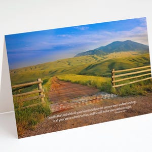 May include: A greeting card featuring a scenic landscape with a dirt road leading towards a mountain. The image includes a wooden fence and a quote from Proverbs 3:5-6. The sky is blue with some clouds, and the hills are green.
