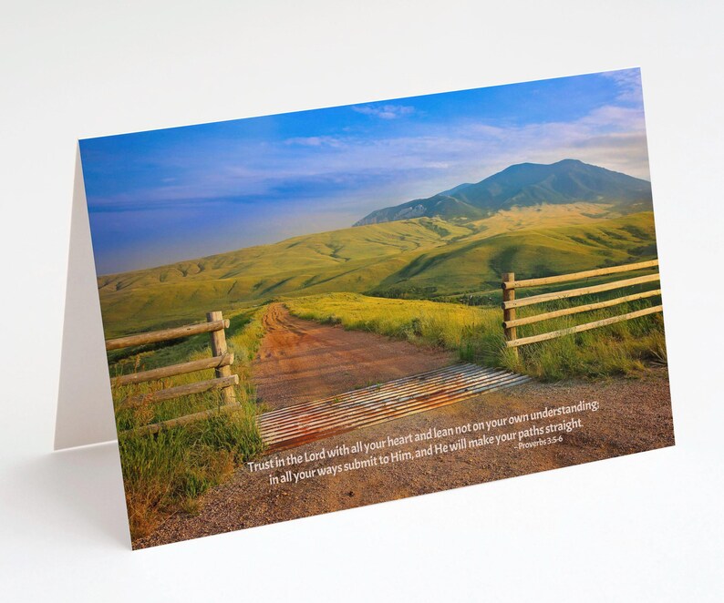 May include: A greeting card featuring a scenic landscape with a dirt road leading towards a mountain. The image includes a wooden fence and a quote from Proverbs 3:5-6. The sky is blue with some clouds, and the hills are green.