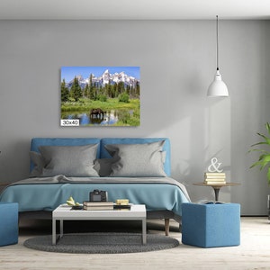 Op de afbeelding: Een canvas print van een bergketen met een bruine eland die in een meer op de voorgrond staat. De print is ingelijst en hangt aan een muur boven een blauwe bank.