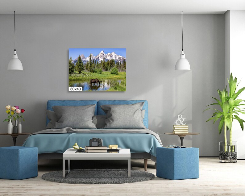 Op de afbeelding: Een canvas print van een bergketen met een bruine eland die in een meer op de voorgrond staat. De print is ingelijst en hangt aan een muur boven een blauwe bank.