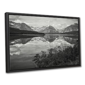 May include: Black and white framed canvas print of a mountain landscape reflected in a lake. The image features snow-capped mountains, a calm lake, and trees along the shoreline. The artwork is encased in a black frame.