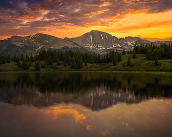 Puede incluir: Un lago de montaña que refleja un cielo de puesta de sol vibrante con una cordillera en el fondo. El lago está rodeado de árboles de hoja perenne y una ladera cubierta de hierba.