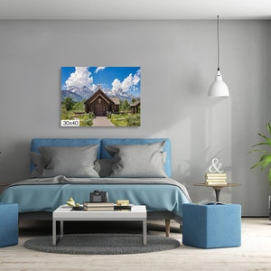 May include: A photograph of a wooden chapel with a cross on the roof, surrounded by green trees and snow-capped mountains in the background. The chapel is located in a field with a path leading to the front door. The sky is blue with white clouds.