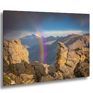 May include: A vibrant landscape photograph featuring a full rainbow arching over a mountain range. The foreground shows rugged, rocky terrain with a view of the mountains under a blue sky with dark clouds.