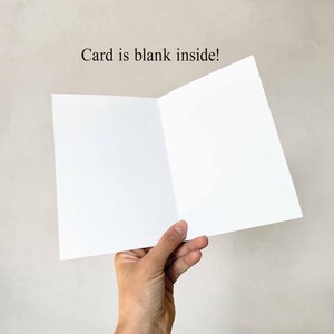May include: A blank white greeting card, held open to display the inside. The card is open, revealing two blank white pages. The text "Card is blank inside!" is printed above the card.