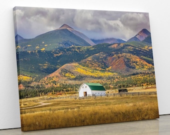 May include: A canvas print of a scenic landscape featuring a white barn with a green roof set against a backdrop of mountains and a cloudy sky. The foreground shows a field of dry grass and a wooden fence.