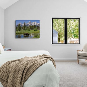 Op de afbeelding: Een slaapkamerinterieur met een wit bed, een bruin gebreid deken, een witte fauteuil en een ingelijst afdruk van een berglandschap met een meer en een eland.