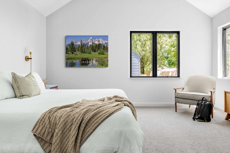 Op de afbeelding: Een slaapkamerinterieur met een wit bed, een bruin gebreid deken, een witte fauteuil en een ingelijst afdruk van een berglandschap met een meer en een eland.