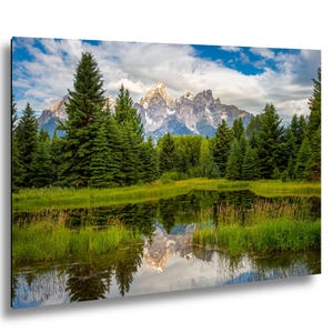 May include: A landscape print showing a mountain range with snow-covered peaks reflected in a calm lake. Green trees and grass surround the water, under a cloudy blue sky. The artwork depicts a tranquil natural scene.