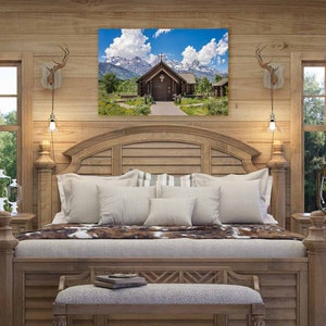 May include: A rustic wooden bedroom with a large bed, two nightstands, and a framed picture of a wooden chapel in the mountains. The bed has a white comforter and a brown and white cowhide throw. The nightstands have drawers and lamps. The picture is above the headboard.