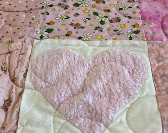 Puede incluir: Un edredón de retazos con un gran corazón rosa aplicado. El edredón presenta varias telas rosas, incluidos estampados florales y de lunares. El corazón es texturizado y el edredón está cosido con un diseño acolchado.