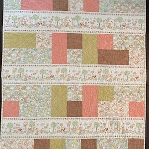 Puede incluir: Una colcha de retazos con un tema de bosque. La colcha está hecha de varias telas en tonos de marrón, verde y rosa. La tela presenta un patrón repetitivo de animales del bosque, árboles y hojas.