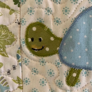 Peut inclure: Gros plan sur une applique de tortue verte et blanche sur un fond de tissu blanc avec un tissu bleu et blanc à pois en arrière-plan.