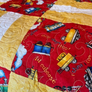 Puede incluir: Colcha de retazos roja y amarilla con temática de tren. La colcha presenta una variedad de vagones de tren, incluyendo un furgón de cola, un vagón de mercancías y una locomotora. La colcha está acolchada con un patrón de espiral y tiene el texto "Lil caboose", "chugga chugga choo!" y "I think I can" en ella.