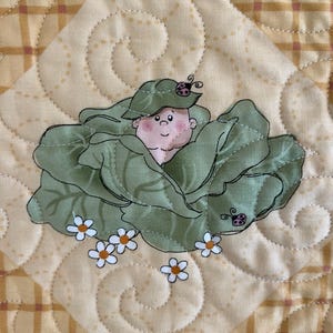 Pode incluir: Um quadrado acolchoado decorativo com um design caprichoso. A imagem central apresenta uma figura infantil aninhada em uma couve verde, com pequenas flores brancas e joaninhas. O fundo é um tecido amarelo claro com um padrão xadrez e redemoinhos acolchoados.