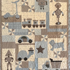 May include: A handmade baby quilt featuring various appliqued shapes in shades of blue, brown, and cream. Designs include a scooter, train, car, turtle, robot, dog, star, and truck. The quilt has a patterned border.
