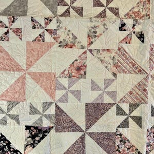 Op de afbeelding: Een quilt gemaakt van verschillende stoffen met bloemen- en geometrische patronen in tinten roze, paars en grijs. De quilt is gemaakt van vierkanten en driehoeken, en de stofpatronen zijn in een herhalend patroon gerangschikt.