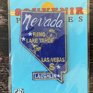Könnte beinhalten: Ein blau-weißer Nevada-Souvenir-Aufnäher in Form des Staates mit den Wörtern "Nevada", "Reno", "Lake Tahoe", "Las Vegas" und "Laughlin". Der Aufnäher befindet sich auf einem hellblauen Hintergrund.