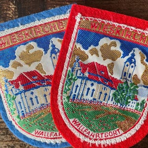 May include: Two embroidered fabric patches featuring a church building with a red roof and a tall tower. The text WIESKIRCHE and WALLFAHRTSORT is stitched in gold and red thread. One patch has a blue felt border, and the other has a red felt border.