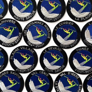 May include: A collection of fourteen circular embroidered patches featuring a skier in yellow clothing on a blue background with white mountains. Each patch displays a specific French ski resort name and elevation in meters, with black borders.