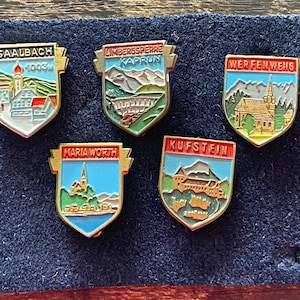 May include: Five enamel pins with colorful images of Austrian towns and landmarks. The pins are gold-colored and have red, white, and blue accents. The towns featured are Saalbach, Limbergsperre Kaprun, Werfenweng, Maria Worth, and Kufstein.