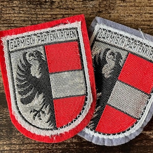 May include: Two embroidered patches with the text "Garmisch-Partenkirchen". Each patch features a shield design with a red and silver color scheme, and a black eagle emblem. One patch has a red border, the other a gray border.
