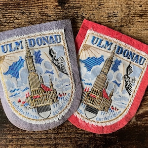 May include: Two shield-shaped embroidered patches, one on a grey felt backing and one on a red felt backing. Each features the Ulm Minster cathedral, the text ULM DONAU, and the text Höchstes Münster der Welt 161m in blue embroidery.
