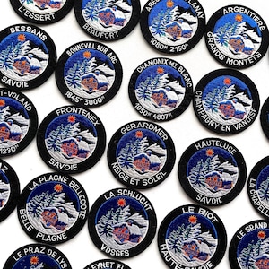 May include: A collection of circular embroidered patches featuring a winter mountain scene with a chalet, snow-covered pine trees, and a bright orange sun. Each patch displays a unique French location name and elevation in white text on a black border.