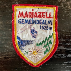 Könnte beinhalten: Gestickter Aufnäher mit rotem Rand, mit dem Text "MARIAZELL GEMEINDEALM" und "1523m". Das Design zeigt eine Berglandschaft mit einer Seilbahn, einem See und einer Blume. Der Aufnäher hat einen weißen Hintergrund mit gelben und grünen Akzenten.