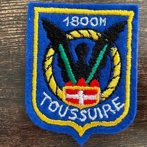 Pode incluir: Um emblema bordado azul com borda amarela apresentando o texto 1800M no topo e TOUSSUIRE na parte inferior. O design inclui uma silhueta de águia preta, um círculo de corda amarelo, detalhes verdes e um emblema de escudo vermelho e branco.