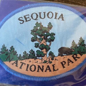 Könnte beinhalten: Ovaler Aufnäher mit der Aufschrift "SEQUOIA NATIONAL PARK". Das Design zeigt einen großen Baum, weitere Bäume und einen Bären vor einem hellblauen Himmel. Der Aufnäher ist blau umrandet. Maße nicht verfügbar.
