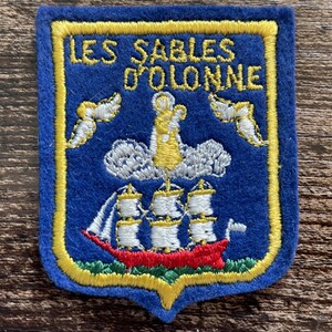 Peut inclure: Un écusson brodé bleu de forme rectangulaire avec une bordure jaune portant le texte LES SABLES D'OLONNE en fil jaune. Le motif représente un voilier blanc à coque rouge, des nuages blancs et deux oiseaux blancs sur fond bleu.
