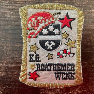 May include: A rectangular embroidered patch with a gold border featuring a red and white carnival hat, a black shield with white stars and crossed hammers, red and gold stars, and the black embroidered text K.G. ROATHEMER WENK on a beige background.