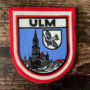 May include: A shield-shaped embroidered patch with a red border and white stitching. The center features a light blue background with a black illustration of the Ulm Minster and a smaller white shield containing a black bird, with the text ULM above.
