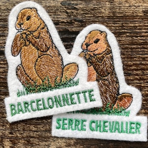 Op de afbeelding: Twee geborduurde patches met bruine marmotten die in groen gras zitten. De linker patch heeft de tekst BARCELONNETTE in het groen en de rechter patch heeft de tekst SERRE CHEVALIER in het groen. Beide hebben een witte vilt achterkant.
