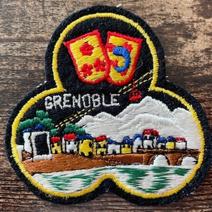 Op de afbeelding: Een zwarte geborduurde patch met een gele rand en de tekst GRENOBLE in het wit. Het ontwerp bevat twee gele schilden met rode en blauwe details, een wit gebergte, een kleine rode kabelbaan en een rij met kleurrijke gebouwen.